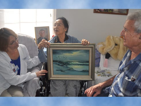 Sara Tyler of Covenant Hospice, left, sits with her client, Masuko Gartman, 82, center, and her husband, Billy Gartman, 81, at Carrington Manor. Masuko holds an original seascape painting that she donated to Carrington.