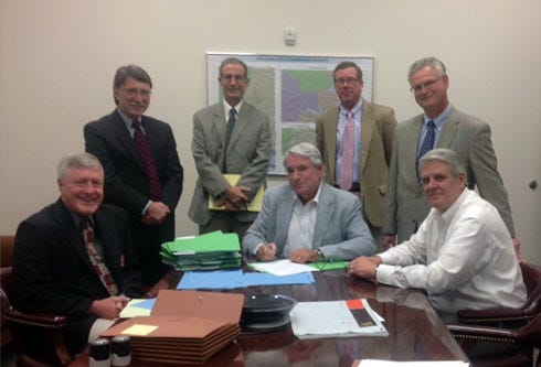 Commissioner Kelly Windes (center) signs the bond financing for the new Okaloosa County Courthouse in CrestviewStanding, from left: Mitchell Owens, RBC Capital Markets, LLC; Mark Mustain, Nabors, Giblin & Nickerson Law Firm; Jody Bear, Crews & Associates; Mark Galvin, First Southwest Company. Seated, from left are Okaloosa Finance Director Gary Stanford, Chairman Windes, and County Administrator John Hofstad.